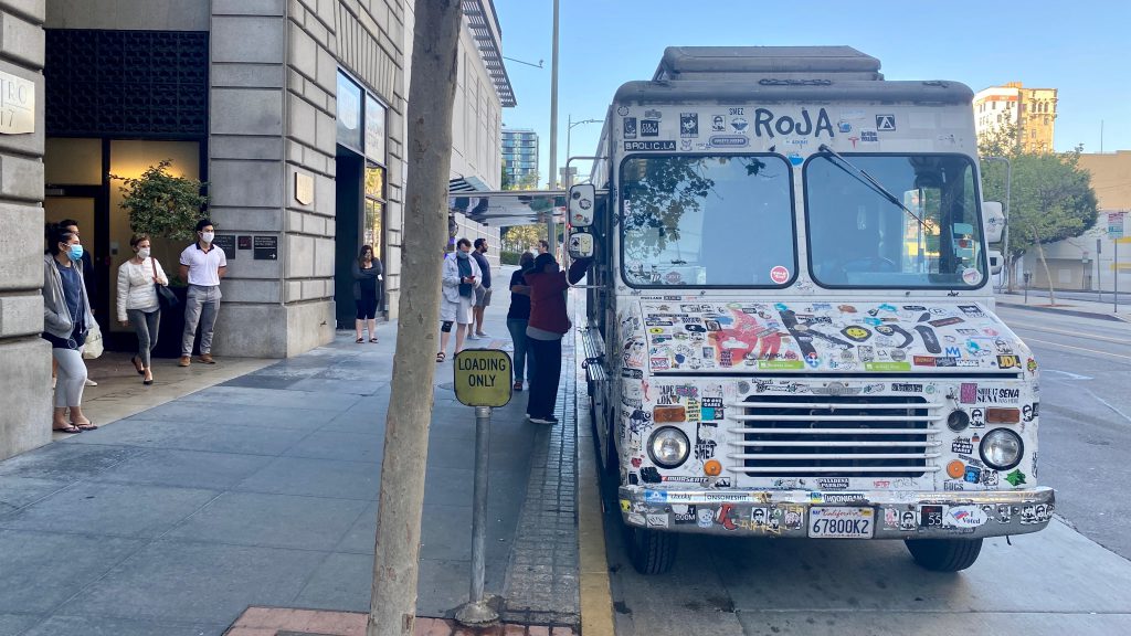 LOS ANGELES, CA, APR 2020: masked residents in Downtown wait for food at sticker-covered Kogi catering truck, parking outside apartments during Covid-19 virus lock down.